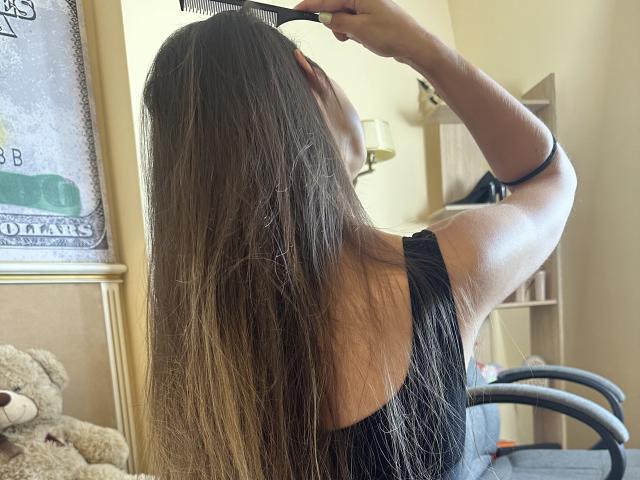 Stefanni with long brown hair, wearing a black sleeveless top, using a comb to detangle her hair, standing in a room with a beige wall and a small teddy bear.