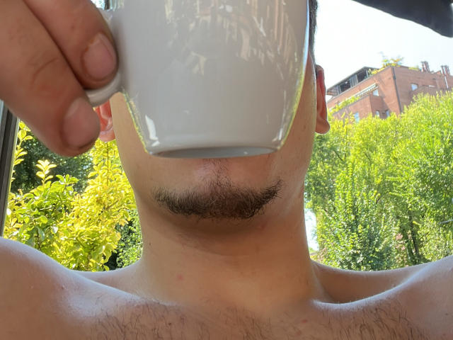 MarkXHot's face is in the foreground, holding a white mug to his mouth, with a mustache and visible neck and shoulder. Greenery and a building are in the background.