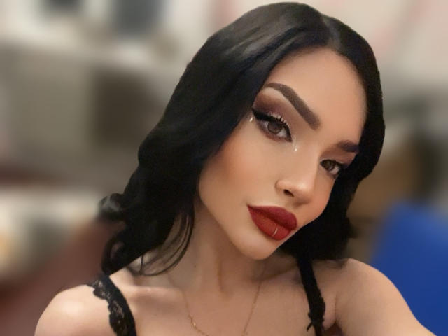 MoiraMilicent has black wavy hair, dark eyebrows, heavy eyeliner, long eyelashes, and red lipstick. She's wearing a black lace top, with a subtle gold necklace.