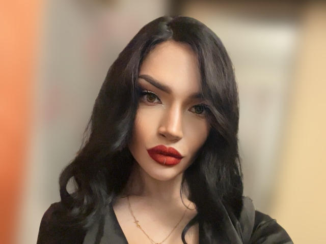 MoiraMilicent has black wavy hair, red lipstick, and a serious expression. She's wearing a black top with a gold necklace, looking straight at the camera.