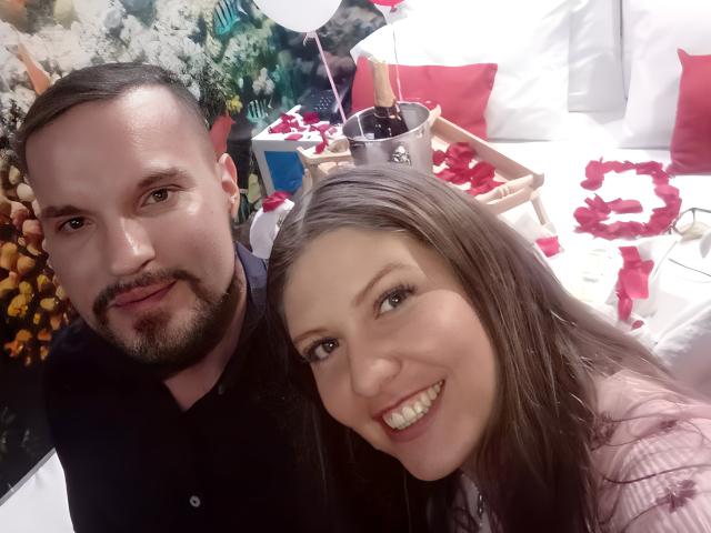 MsMiaMrSergio with a beard and MsMiaMrSergio with long brown hair, smiling, in a cozy room with white pillows and red rose petals, holding a drink.