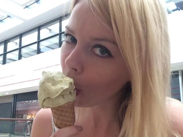 NicolleLinharts-hot with blonde hair, sucking on a vanilla ice cream cone in a mall, eyes half-open, looking cute, wearing a white top, holding the cone with her right hand.