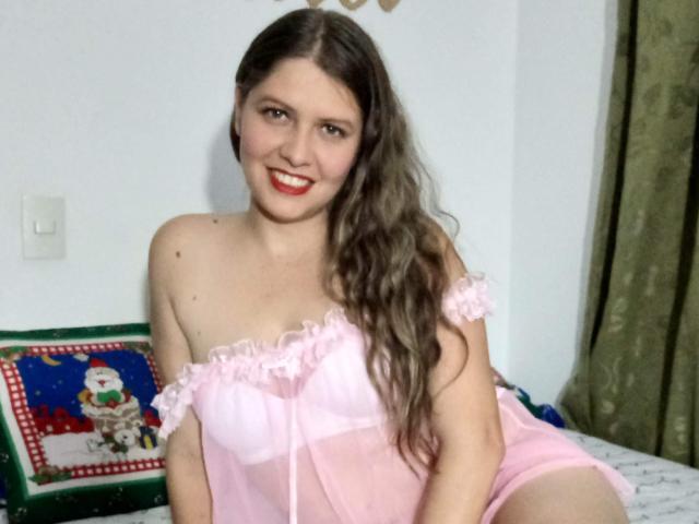 MsMiaMrSergio with long brown hair, smiling, wearing a pink sheer babydoll, showing her small tits and bare ass, sitting on a bed with green and red pillow.