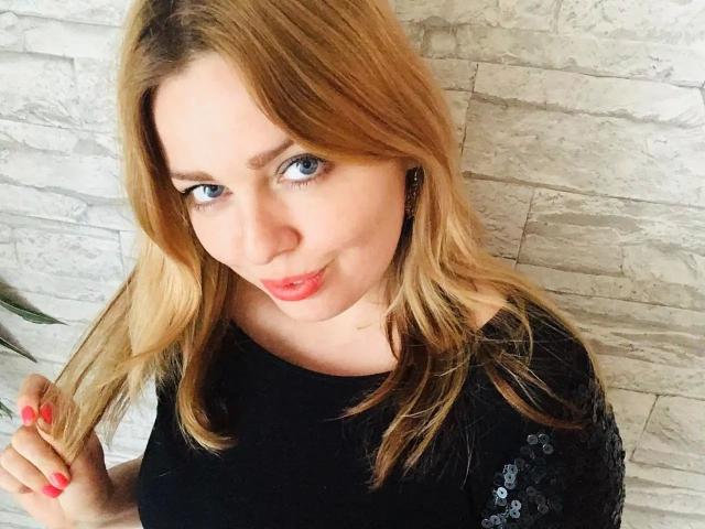 NicolleLinharts-hot has blonde hair, blue eyes, and red lipstick, wearing a black sequined top, with her right hand holding a lock of hair, pink nails.