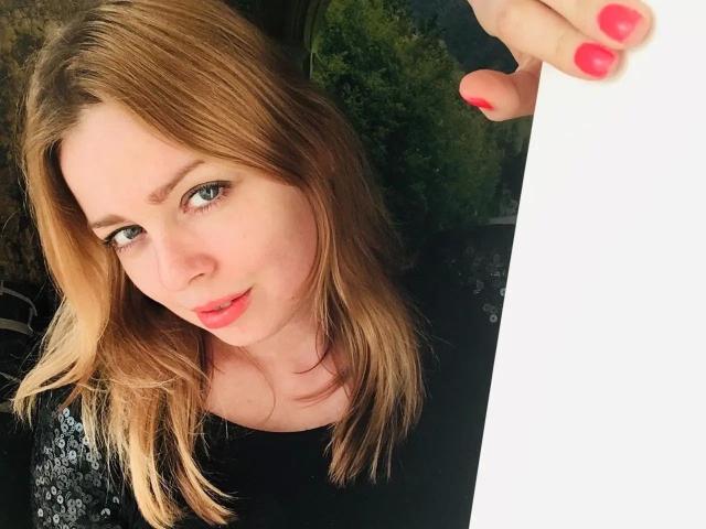 NicolleLinharts-hot with blonde hair, green eyes, pink lipstick, black top, holding white paper, pink nails, slight smile, looking up.