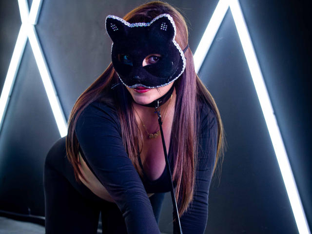 ZoeFerran with long brown hair, black cat mask, black long-sleeve top, and black pants, bending forward, showing her tits, wearing a black choker, in a dark, neon-lit room.