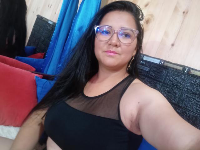 Philipa with black hair, glasses, pink lipstick, and black sheer top showing her tits, standing in a room with blue curtains and wood walls.