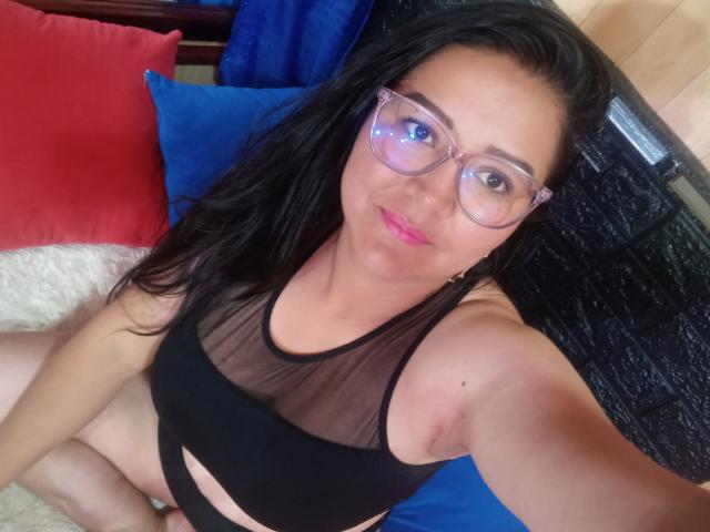 Philipa with black hair, glasses, and pink lipstick, wearing a sheer black top, sitting with legs crossed, showing her bare arms and a small mole on her right arm.
