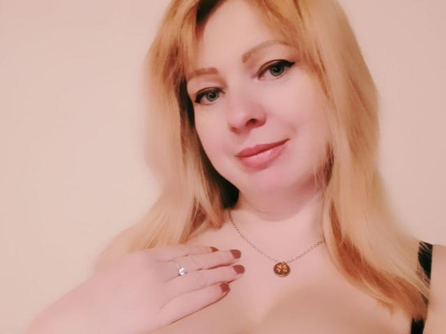 DoriaRay has blonde hair, green eyes, and is smiling with pink lipstick. She's touching her chest with a hand showing a ring, wearing a necklace with a round pendant.