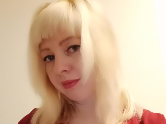 DoriaRay with blonde pigtails, pale skin, red lipstick, and blue eyes, wearing a red shirt, has a slight smile. Her tits are not visible.