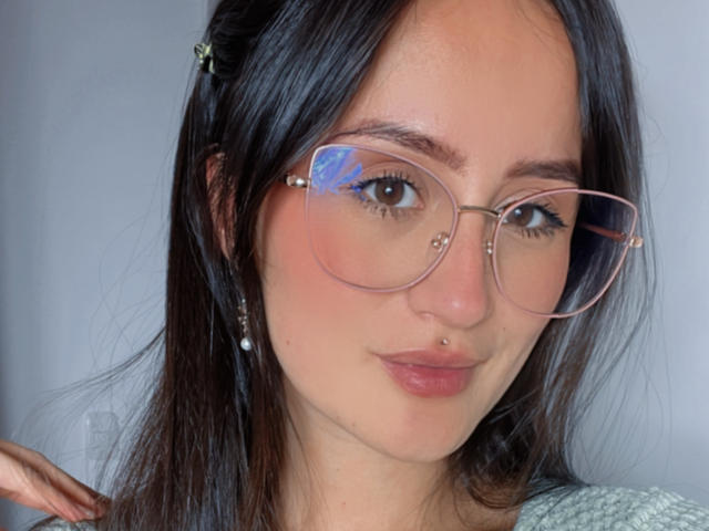 NinaBryons has dark hair, a nose ring, and is wearing oversized pink glasses. She's sporting a subtle pink lip color and a green patterned shirt. Her skin is fair.