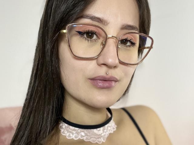 NinaBryons with brown glasses, pink eyeshadow, black choker, and pink lace necklace, has a nose piercing and pink lipstick, showing her face and straight brown hair.