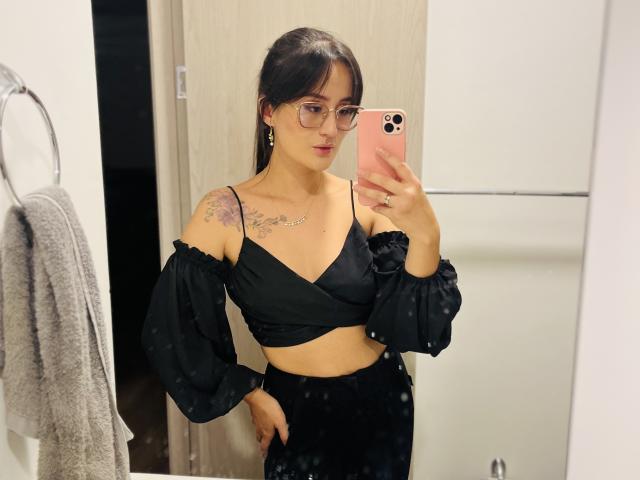 NinaBryons with glasses, black off-shoulder top, and black pants, showing her tits and a tattoo on her left shoulder, taking a mirror selfie with a pink phone.