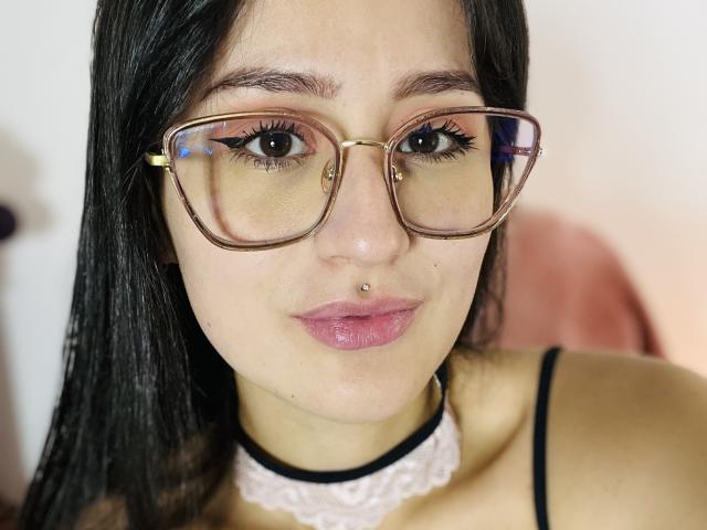 NinaBryons with big brown eyes, wearing copper glasses, black choker, lace necklace, and pink lipstick, has a nose ring and a small lip piercing, black hair, and a serious look.