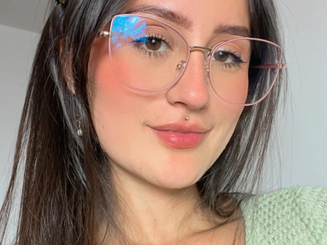 NinaBryons with pink glasses, nose piercing, and pink lips, brown hair, green sweater, subtle smile, reflection of blue light on glasses, soft pink blush on cheeks.