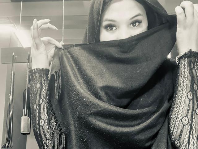 MalakCote wearing a black lace top with see-through patterns, black mask covering her nose and mouth, black headscarf, and brown eyes looking at the camera. Red-orange wall background.