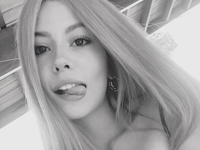 GalliBarbara, com cabelo longo e liso, língua para fora, olhando para a câmera, usando brincos, foto em escala de cinza, mostrando seu rosto, boca ligeiramente aberta, com uma expressão divertida e sensual..