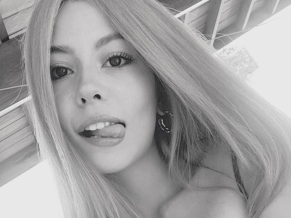 GalliBarbara, com cabelo longo e liso, língua para fora, olhando para a câmera, usando brincos, foto em escala de cinza, mostrando seu rosto, boca ligeiramente aberta, com uma expressão divertida e sensual..