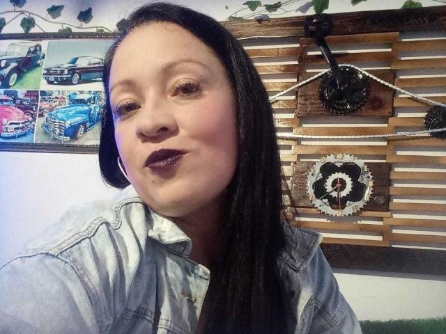CharloteEzra with dark lipstick, slightly parted lips, and long black hair, wearing a light denim jacket, smiling in a selfie, showing her round face and small nose.