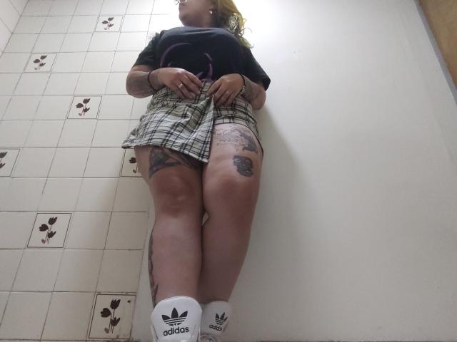 GineBralkino stands with legs together, wearing a plaid skirt, black shirt, tattoos on thighs, and Adidas socks, showing her ass and tattoos, taken from a low angle.