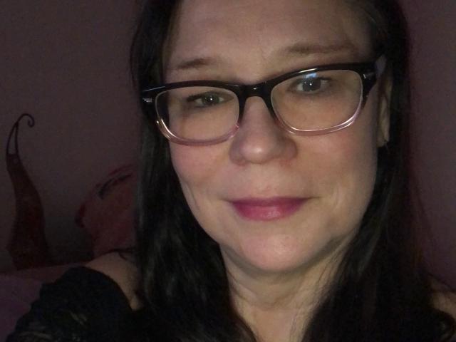 MarylouMar has pale skin, dark brown hair, and is wearing black glasses with a pinkish nose and pink lipstick. Her tits are not visible in the image.