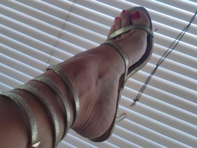 Rosyta's foot in a gold strappy high heel with pink-painted toes, showing her ankle and sole, angled upwards, against white blinds.