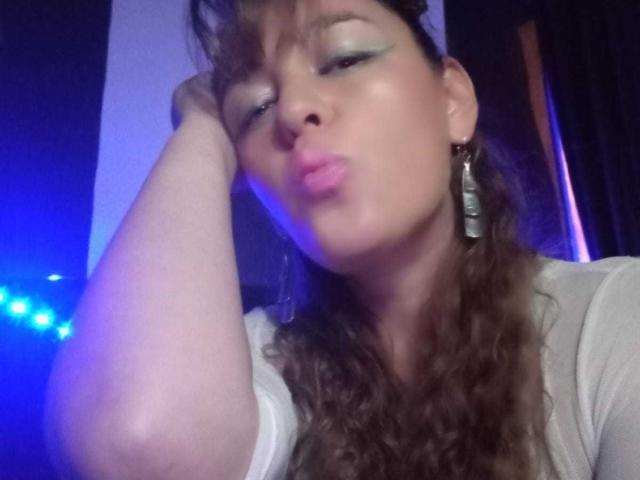 AmandaHotL with pouty lips, blue eyeshadow, curly brown hair, white shirt, and earrings, resting her head on her hand, looking cute. Blue light in the background.