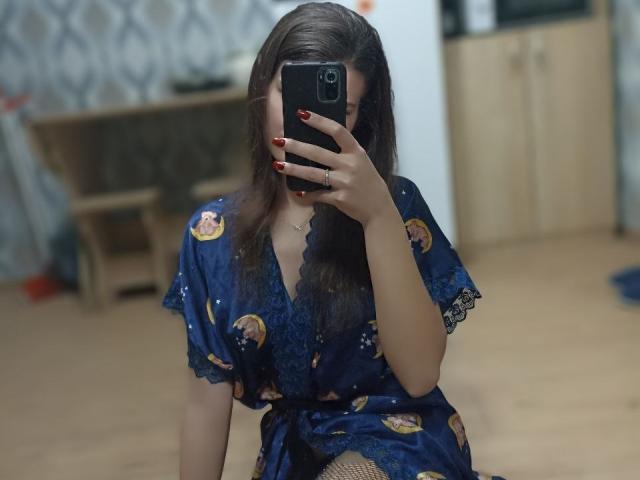 LauraLee69 with long brown hair, wearing a blue floral dress, sitting, holding a phone, covering her face, red nails, wooden floor, wooden cabinet, patterned wallpaper, blurry background.
