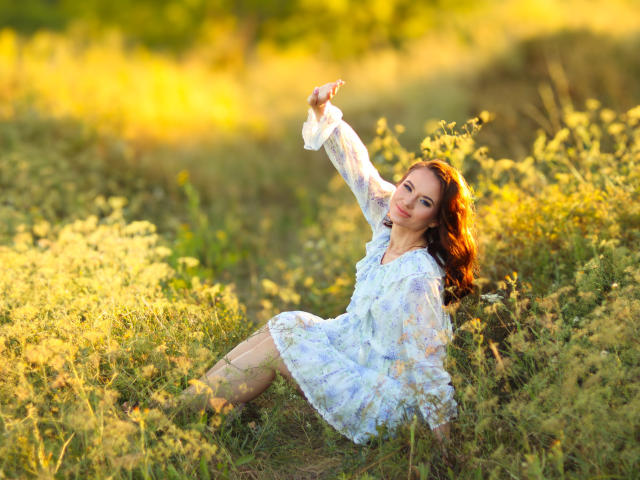 KaraCriegs mit roten Haaren, in einem weißen Blumenkleid, sitzt auf einem sonnigen Feld, hält ein weißes Tuch über ihrem Kopf, lächelt, und ihr Hintern ist teilweise von Gras verdeckt..