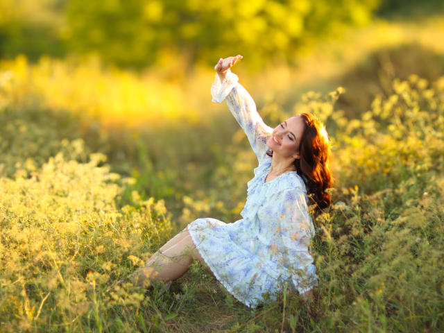 KaraCriegs with red hair, wearing a floral dress, sitting in grass, smiling, arms raised, sunlight on her face, looking happy, in a field of yellow-green plants.