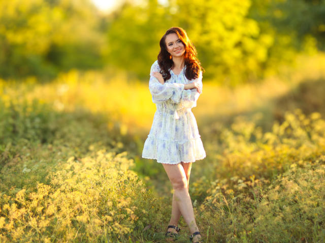 KaraCriegs standing in a sunlit field, wearing a white lace dress, arms crossed, smiling, brown wavy hair, green eyes, bare legs, black sandals, soft golden light.