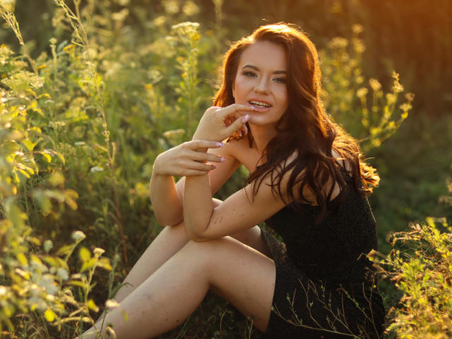 KaraCriegs with long brown hair, wearing a black sparkly dress, sitting in sunlit grass, pointing at her smiling mouth, showing off her bare thighs and cute ass.