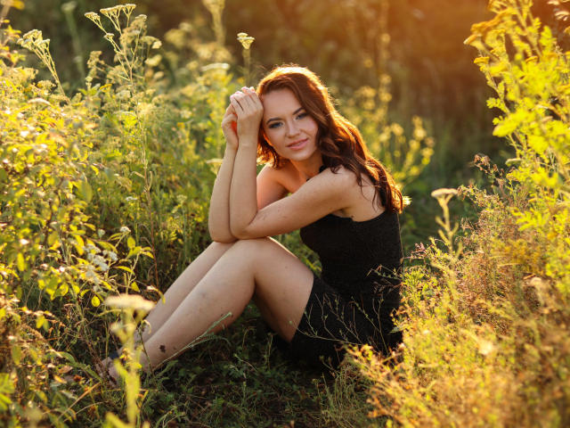 KaraCriegs with wavy red hair, black dress, sitting in grass, legs crossed, hands on head, smiling, sunlight, green foliage, warm glow, pretty face, cute, relaxed, natural.