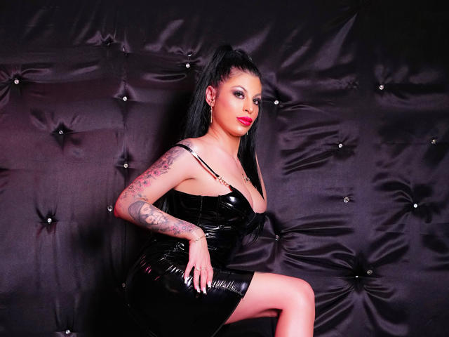 AmmyMiss met tatoeages, in een zwarte latexjurk en met een hoge paardenstaart, poseert met haar hand op haar knie, waarbij haar glanzende zwarte jurk en rode lippenstift te zien zijn, voor een zwarte, gestructureerde achtergrond..