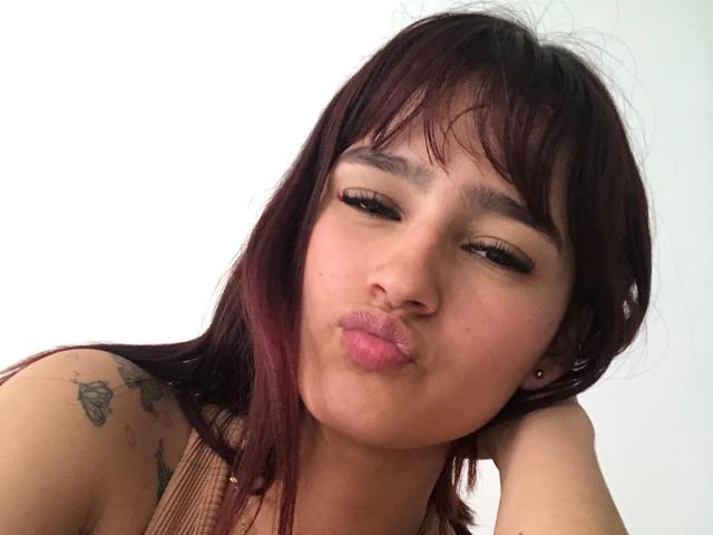 MelodyMansson has puffy pink lips, dark brown hair with red highlights, and a tattoo on her right shoulder. She's making a pouty face with her eyes half-closed.