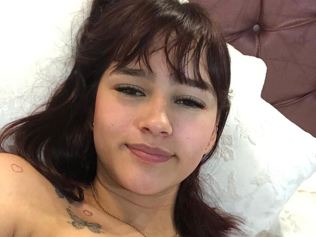 Melody Mansson tiene una piel marrón media, cabello rojo oscuro y está sonriendo con los ojos medio cerrados. Tiene tatuajes en el hombro y el pecho, y lleva un collar de oro..