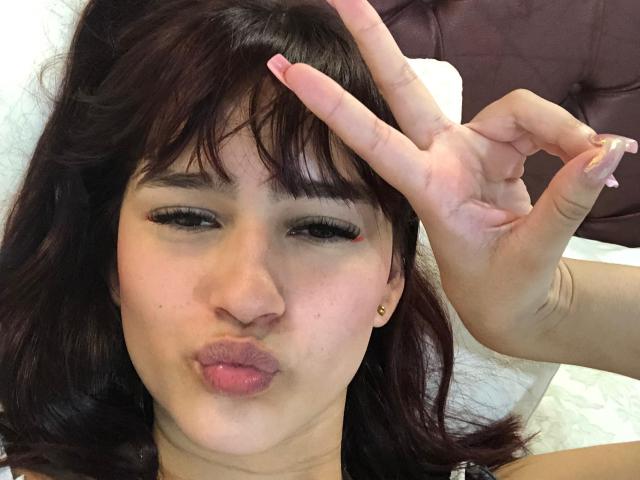 MelodyMansson with dark brown hair, pouty lips, and a red scratch above her right eye, making a peace sign with her right hand, showing off her long, pink nails.