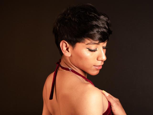 Anddyys with short black hair, closed eyes, and shiny skin, wearing a red halter top, holds her right breast with her left hand, against a dark background.