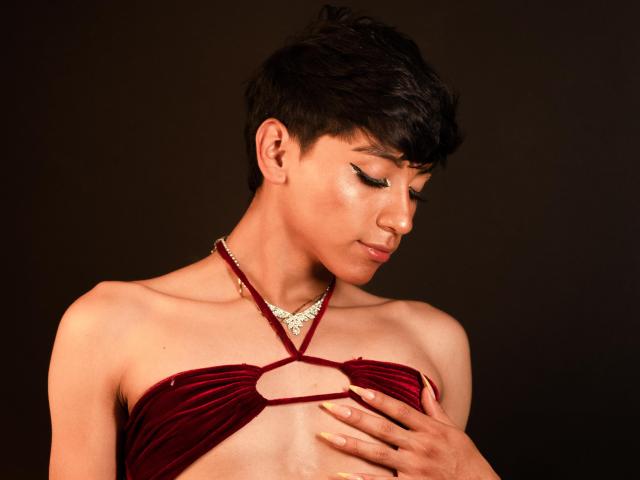 Anddyys has short dark hair, wearing a red velvet top that barely covers their small tits, with a hand on their chest, looking down, against a dark background.