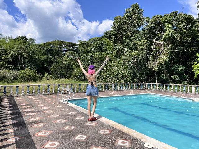 SarahRousen with arms raised, wearing a pink headscarf, denim shorts, and flip-flops, standing by a blue pool, back to the camera, on a patterned tile deck.