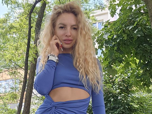 LoreenBlanc with long, wavy blonde hair, wearing a blue cut-out top, standing in a green, leafy area, holding her chin, showing a small, sexy, cute smile.
