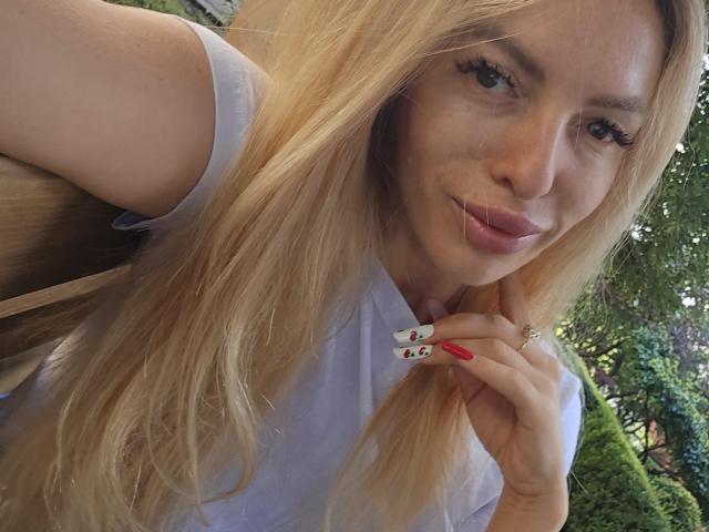 LoreenBlanc with long blonde hair, pouty pink lips, wearing a white shirt, holding a finger with red nails and a black/white nail, looking at the camera.
