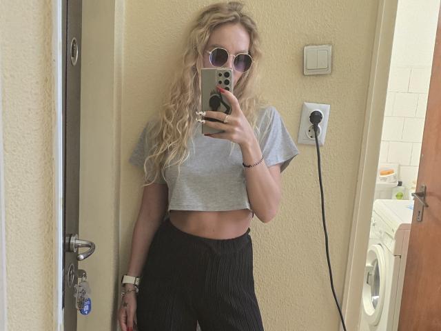 LoreenBlanc, met krullend blond haar, draagt een grijze crop top en zwarte broek, ze houdt een telefoon vast, bedekt haar borsten, draagt roze zonnebril en staat in een badkamer..