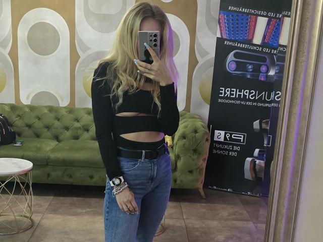 LoreenBlanc with blonde hair, black long-sleeve crop top, blue jeans, holding phone, mirror selfie, green tufted couch, standing, white wall with circular patterns, beige floor.