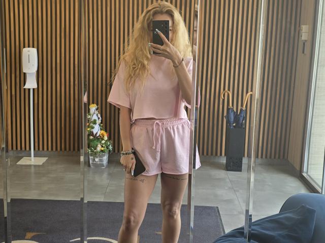 LoreenBlanc, com cabelo loiro, vestindo shorts e blusa rosa, segurando o celular em um selfie no espelho, com tatuagens visíveis na coxa, uma pulseira rosa, em pé em um quarto moderno, com uma cadeira azul em primeiro plano..