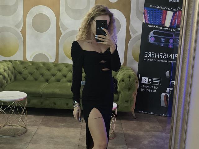 LoreenBlanc mit lockigem, blondem Haar, trägt ein schwarzes, langärmliges Kleid mit Ausschnitten und einem Schlitz am Oberschenkel, hält ein Telefon, Selfiemotiv im Spiegel, im Hintergrund ein grünes, gepolstertes Sofa..