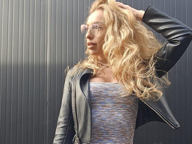 LoreenBlanc, con i capelli biondi mossi, indossa una giacca di pelle nera e una camicetta a quadri, con una mano tra i capelli, occhiali chiari, guarda di lato, con sullo sfondo un muro scuro a righe verticali..