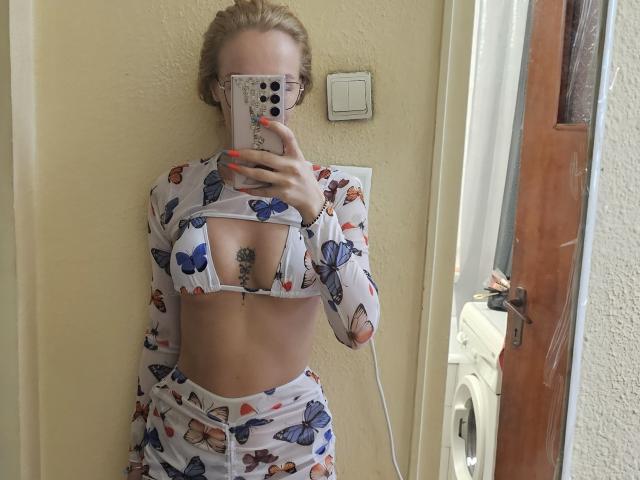 LoreenBlanc, con cabello rubio, gafas y ropa interior con estampado de mariposas, mostrando sus senos en un selfie frente a un espejo, sosteniendo un teléfono con una funda blanca, uñas anaranjadas y un tatuaje..