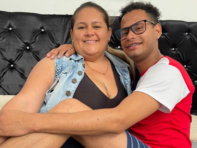 AntonioAndMichel with medium tits in a black bra, sitting next to AntonioAndMichel in a red and white shirt, arms around each other, smiling on a black tufted couch.