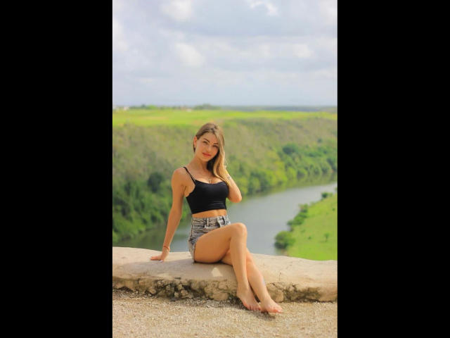 BerlinerGirl89-hot sits on a stone ledge, wearing a black crop top and denim shorts, showing her tits and ass, with her legs crossed, barefoot, and blonde hair.